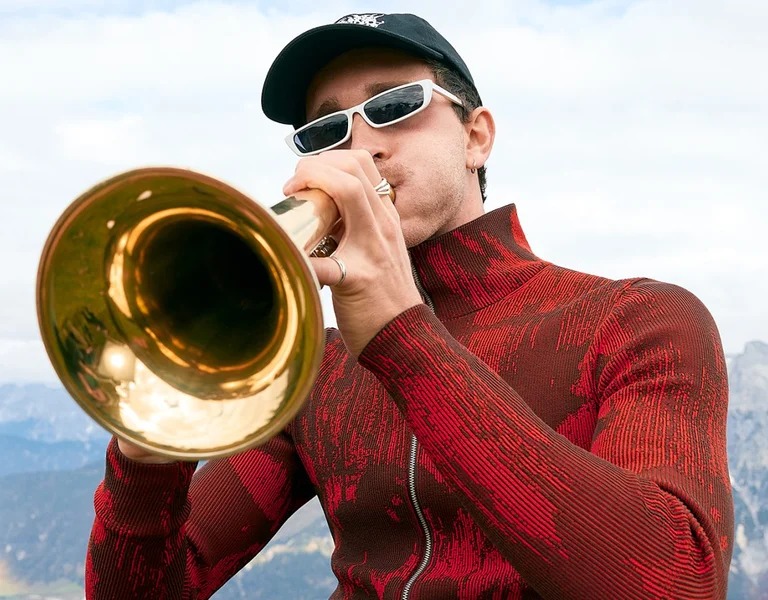 Modern musician in sunglasses and a red jacket playing a trumpet against a background of mountains and cloudy sky.