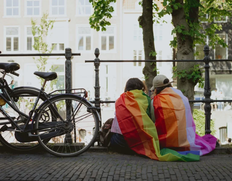 Two people draped in a rainbow Pride flag sitting by a canal, watching the Canal Parade in Utrecht, Netherlands, a major LGBTQ+ travel destination for 2026.