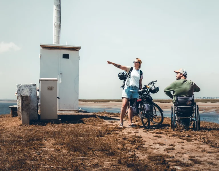 Two people with bikes by a small lighthouse; one points toward the water.