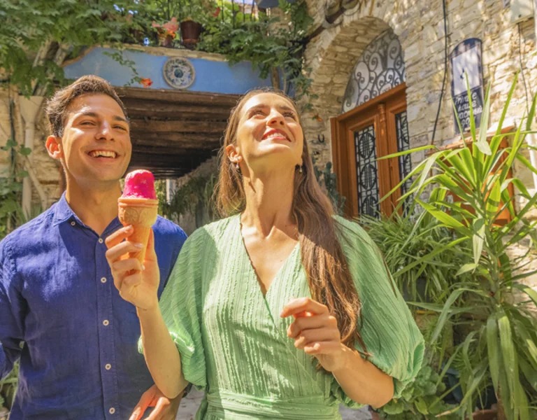 Smiling man and woman eating ice cream outdoors by a stone building with lush greenery.