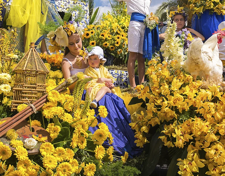 People on a parade float covered with bright yellow flowers under a blue sky.