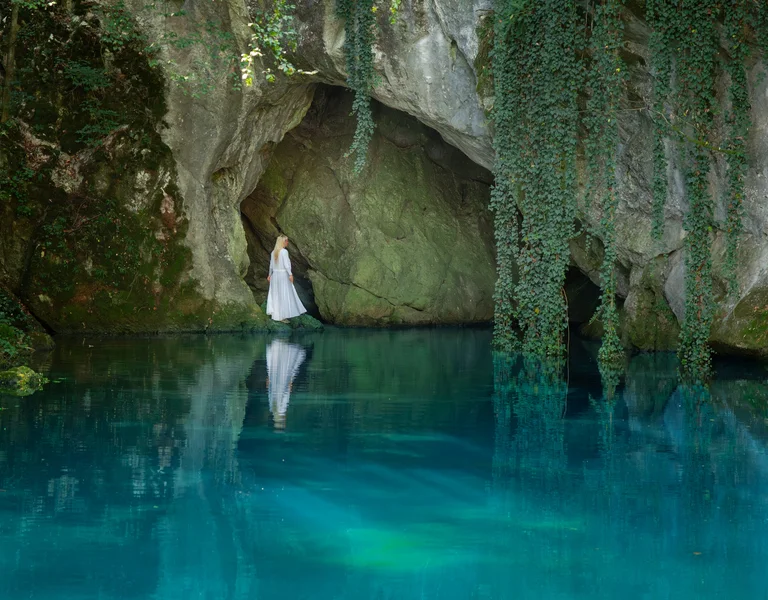 Woman in white dress stands by a cave entrance above clear blue water, surrounded by greenery.