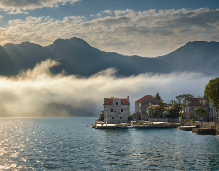 Coastal village with stone houses, mist over water, and mountains in the background.