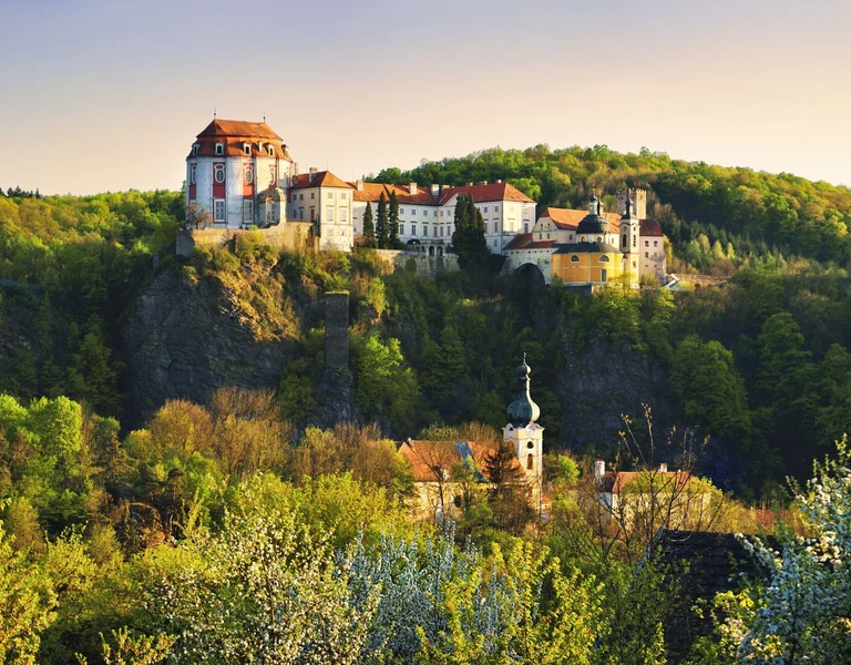 Castle complex atop a forested hill in golden sunlight, with a church below.