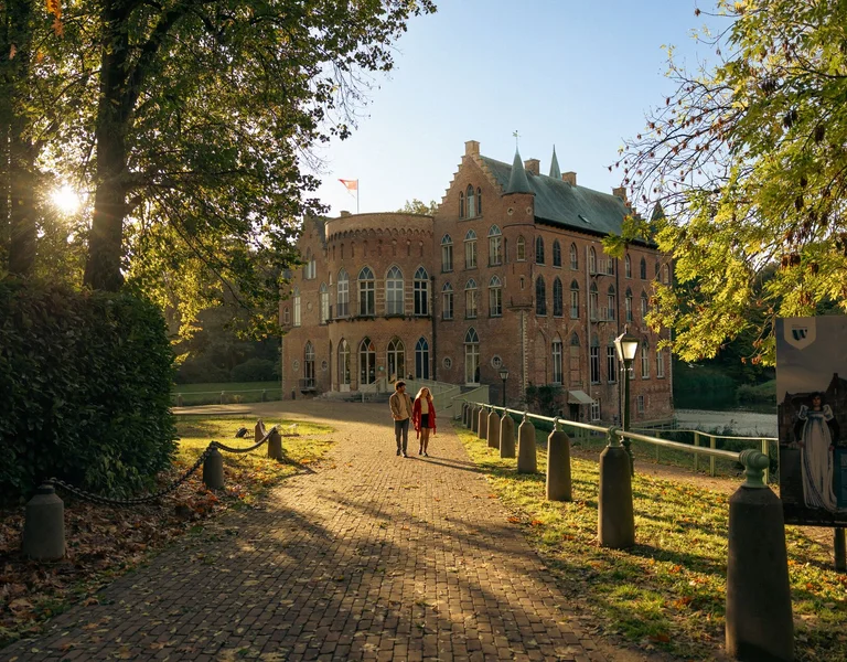 Two people walk on a tree-lined path toward a brick mansion in warm sunlight.