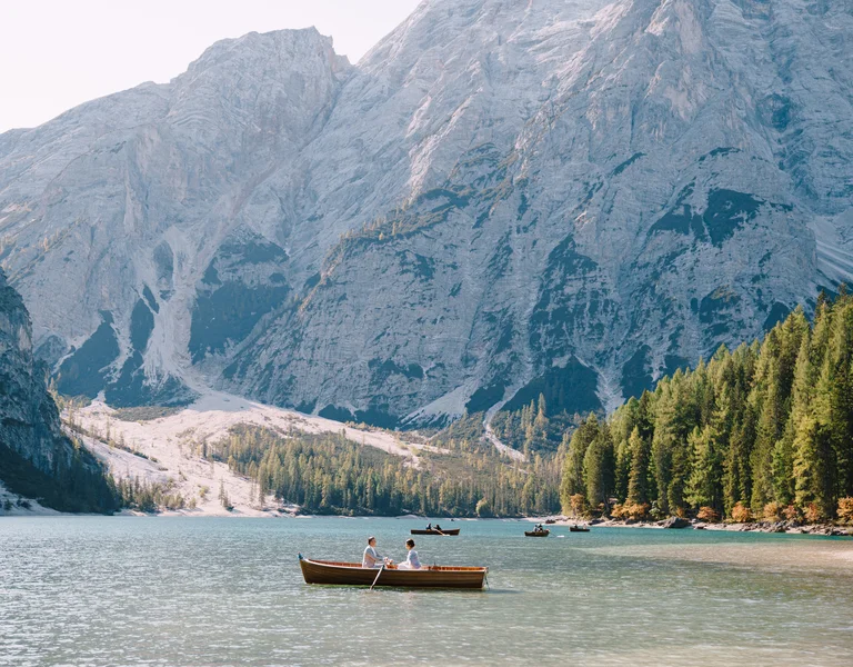 Two people in a wooden boat on a lake surrounded by pine trees and tall mountains.