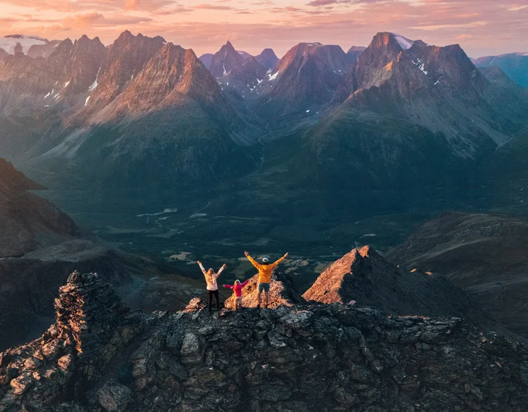 Three people celebrate on a mountain peak at sunset with dramatic mountains behind them.