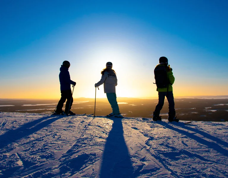 Three people with ski poles standing on snowy hill at sunset.