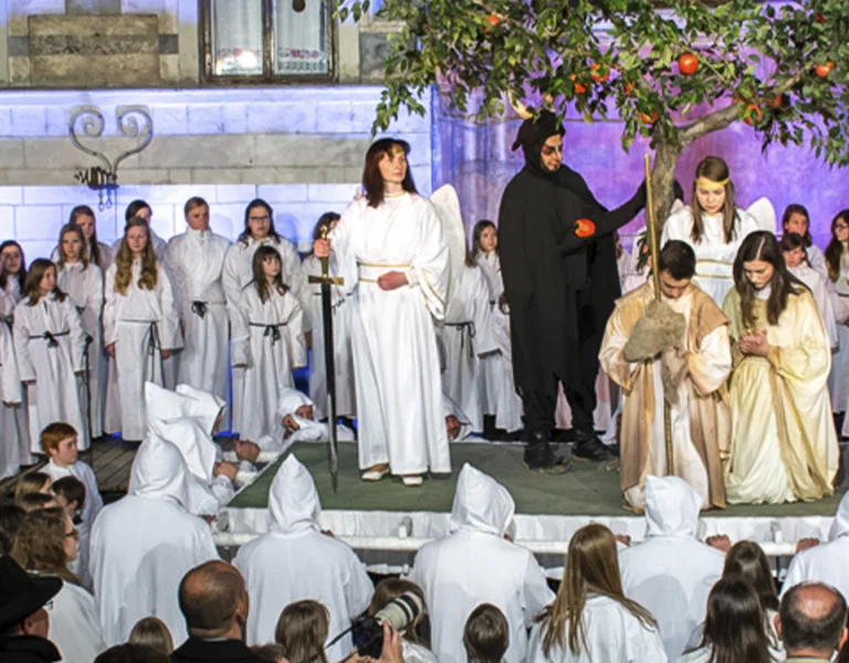 Large group of people in white robes performs a play on stage; one figure wears a bright red robe.
