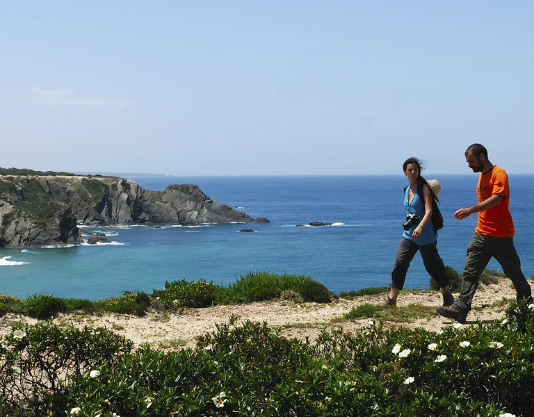 Two people hiking along a coastal cliff with ocean and blue sky in the background.