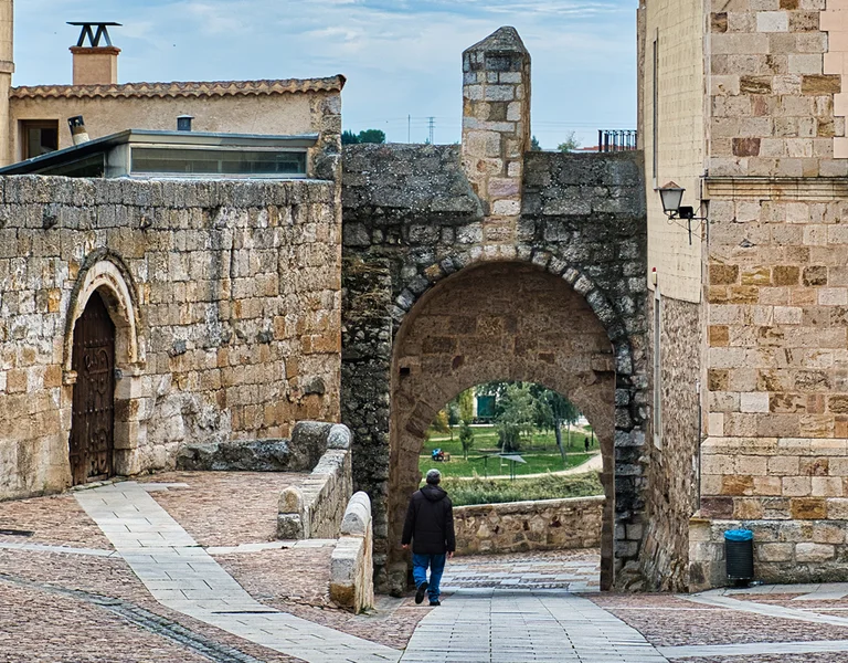Person walks through an old stone archway in a historic, cobblestone plaza.