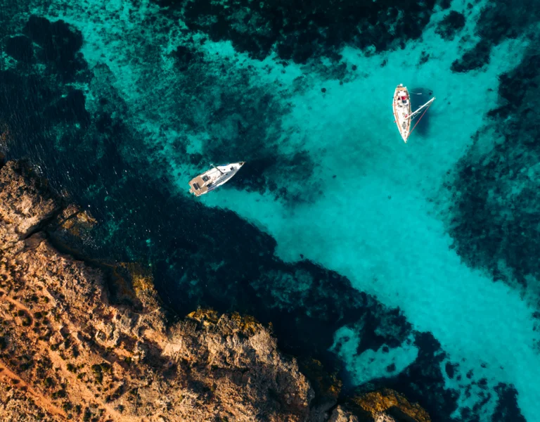 Two sailboats in clear turquoise water near a rocky coastline, photographed from above.