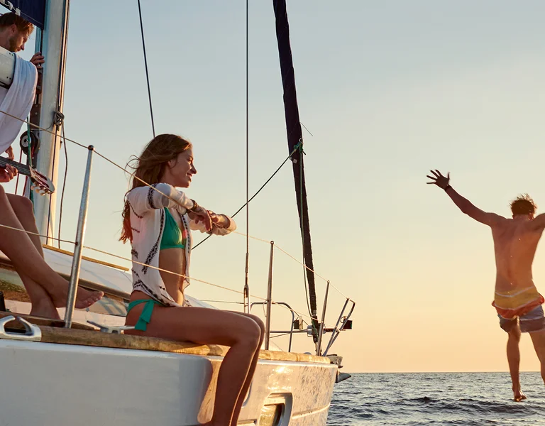 People relaxing on a sailboat while one person jumps into the sea at sunset.