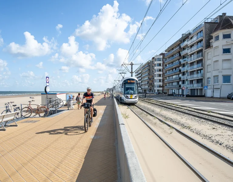 Cyclist rides by the beach near tram tracks and seaside apartments under a blue sky.
