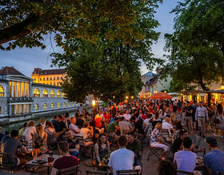 Crowded riverside cafe at dusk with people socializing under trees and glowing lights.