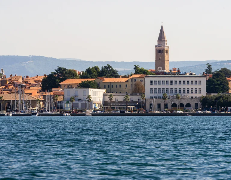 Coastal town with a tall clock tower, waterfront, and mountains in the background.