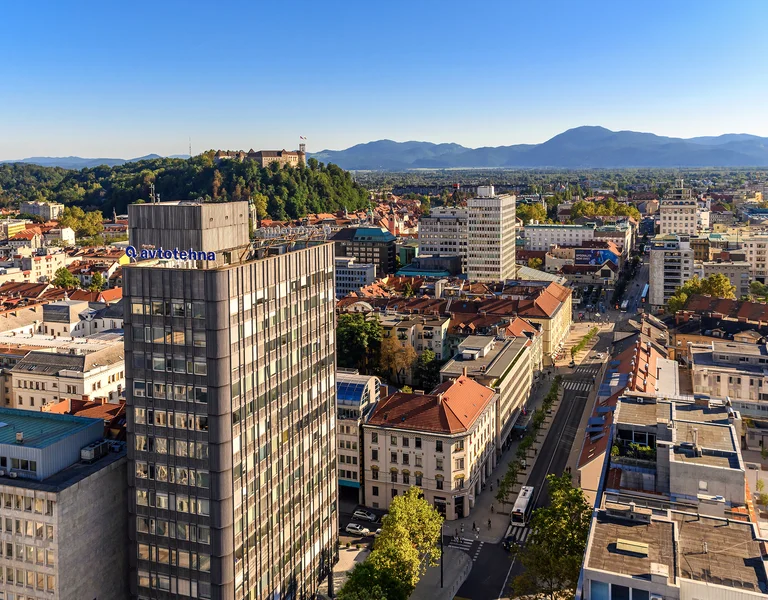 Cityscape with modern buildings, a hilltop castle, and mountains in the background.