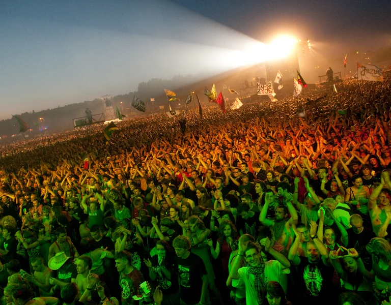 Large crowd at an outdoor concert, hands raised, with bright stage lights at dusk.