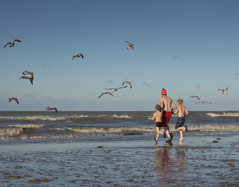 Three children run on a sandy beach with seagulls flying above the waves.