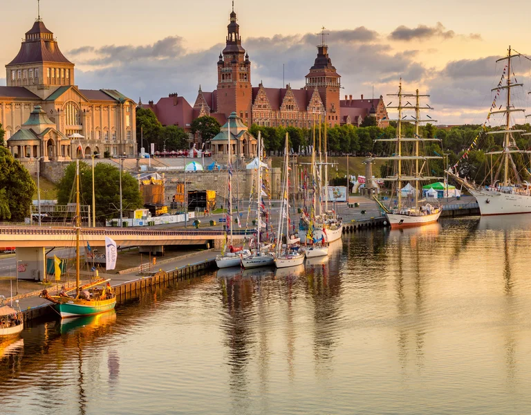 Harbor with sailboats and historic buildings at sunset, calm water reflecting the sky.