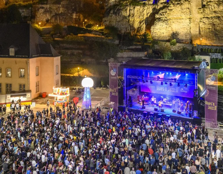 Large crowd at an outdoor concert in a courtyard with stage lights and historic buildings.