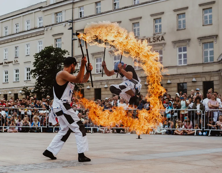 Performer jumping through a flaming hoop held by another person in a city square.