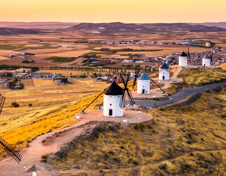 Row of old white windmills on a hilltop at sunset, overlooking a vast rural landscape.