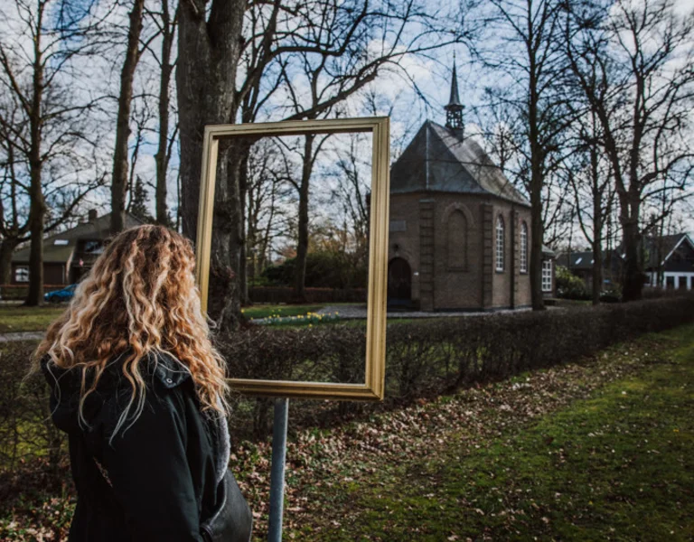 Woman with curly hair looks through an empty frame at a chapel surrounded by bare trees.