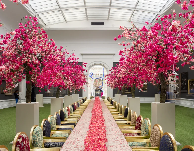 Long banquet table with pink flower petals, flanked by blooming pink trees and ornate chairs.