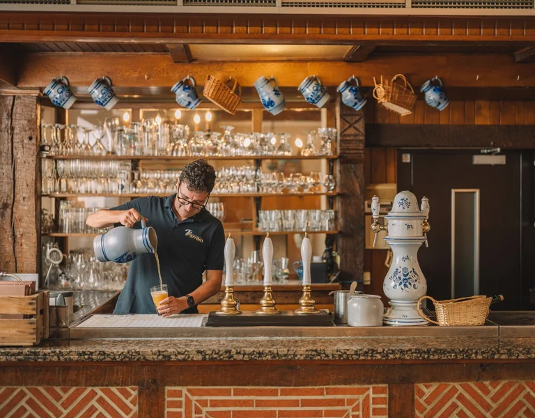 Bartender pouring a drink at a rustic bar with glassware and ceramic mugs on display.