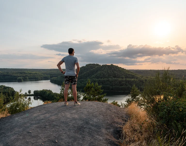 Person standing on a hill overlooking a river and forest at sunset.