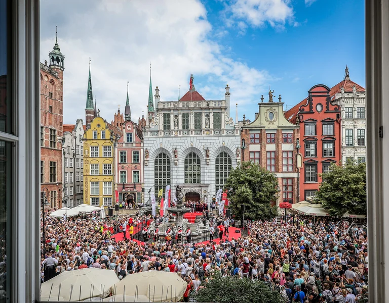 Crowd gathered in a colorful European square framed by open window.