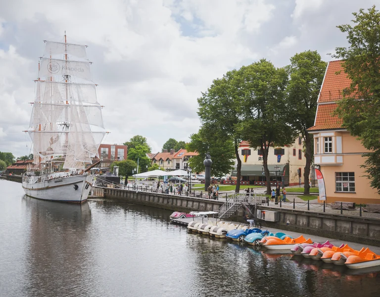Tall ship docked on a riverside with colorful paddle boats and historic buildings nearby.