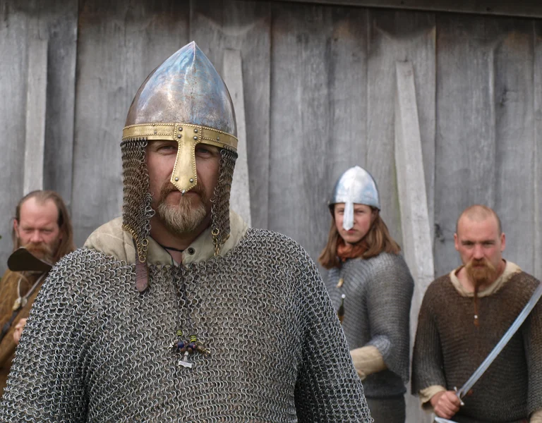 Four people in medieval armor and chainmail, posed with swords against a wooden wall.