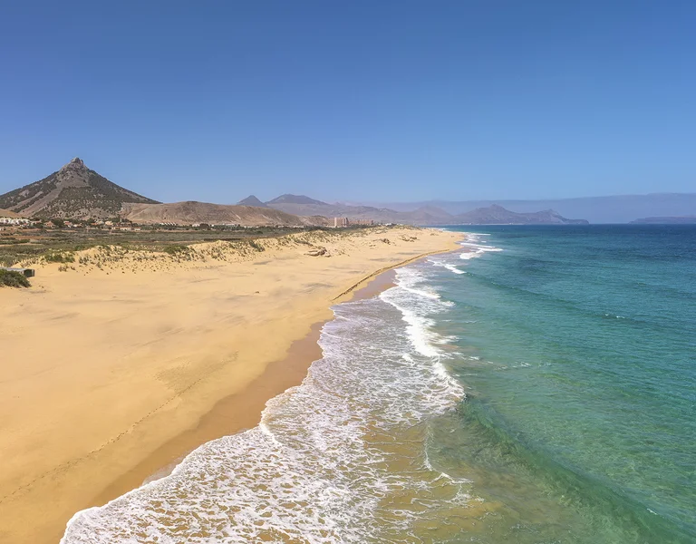 Wide sandy beach with turquoise waves, clear sky, and distant mountains.