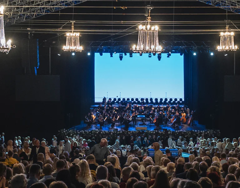 Large audience seated in a theater, facing a brightly lit stage with chandeliers overhead.