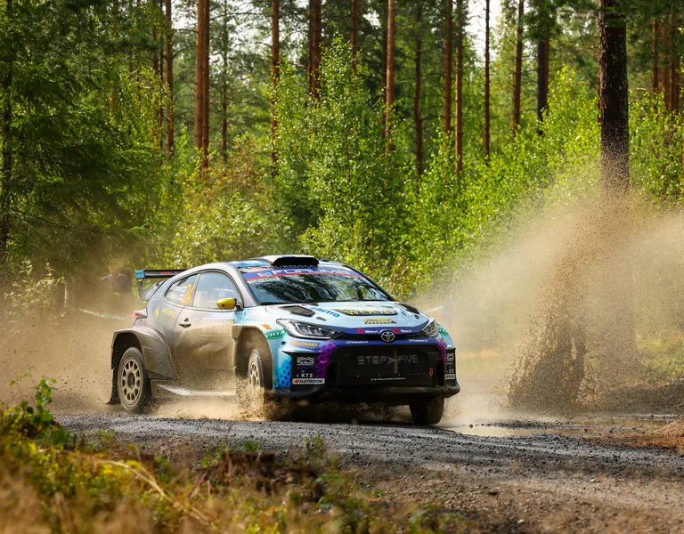 Rally car speeding on a gravel road in a forest, kicking up dust.
