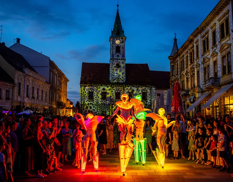 Street performers on stilts lit by colorful lights, surrounded by a crowd at dusk.