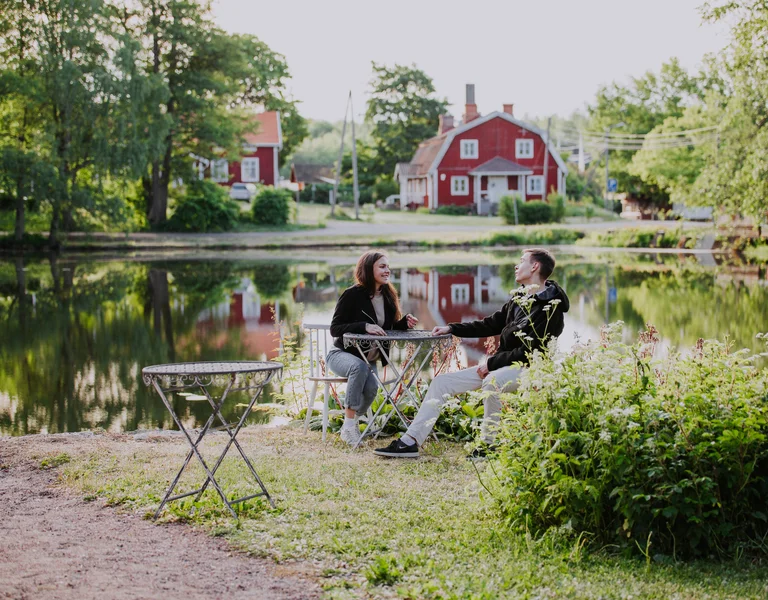 Two people sit at a small table by a pond with red houses in the background.