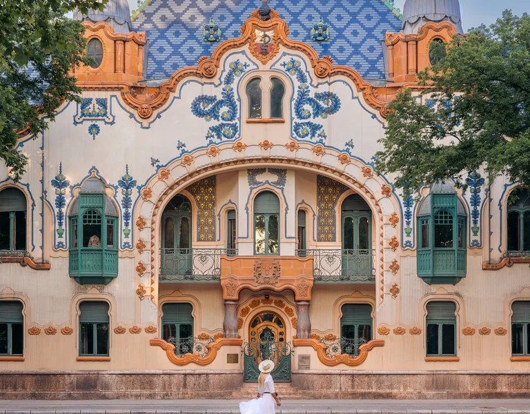 Woman in white dress crossing street in front of ornate, colorful building with arched entry.