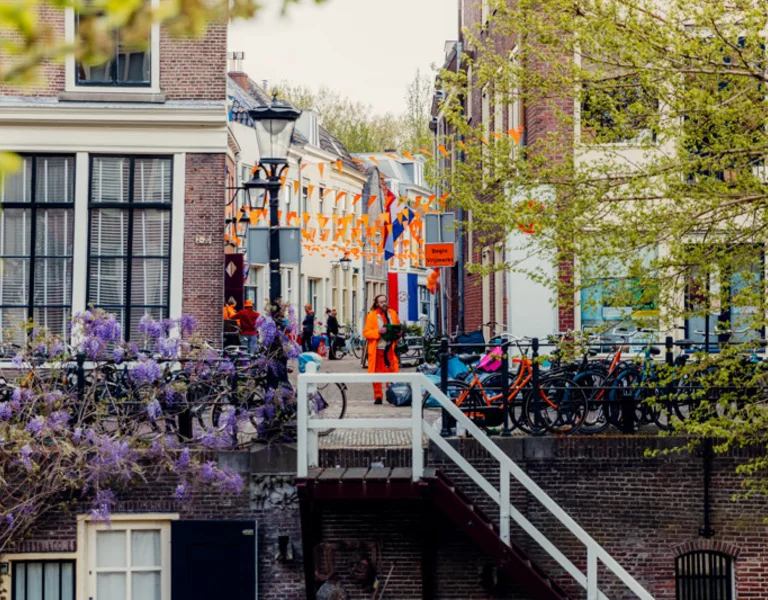 City street decorated with orange flags, people walking, and bicycles by canal railing.