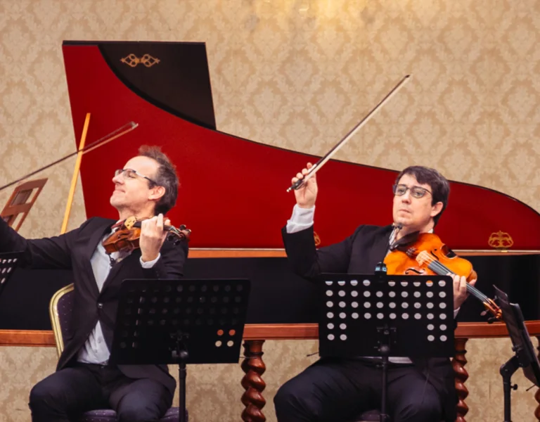 Classical musicians performing with string instruments in front of a red harpsichord.