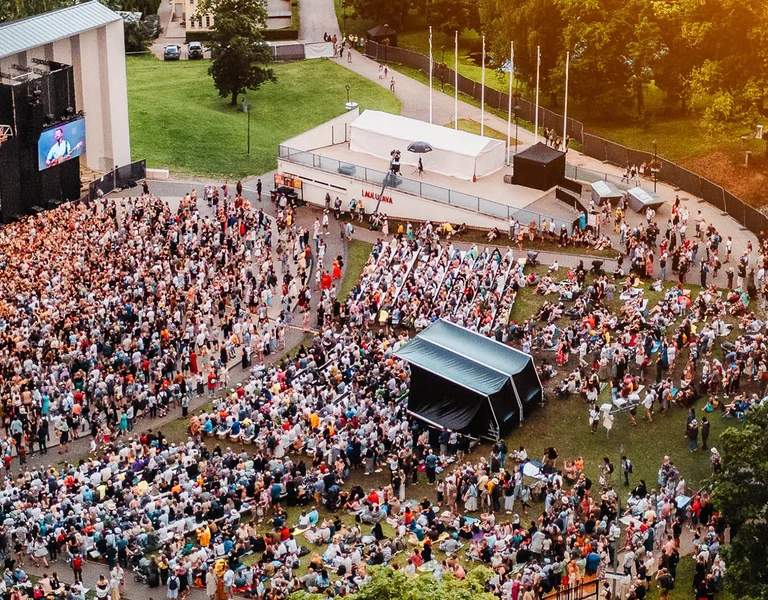 Large crowd gathered at an outdoor concert surrounded by trees at sunset.