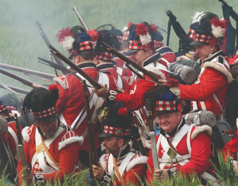 Soldiers in red uniforms with muskets gathered in tall grass during a reenactment.