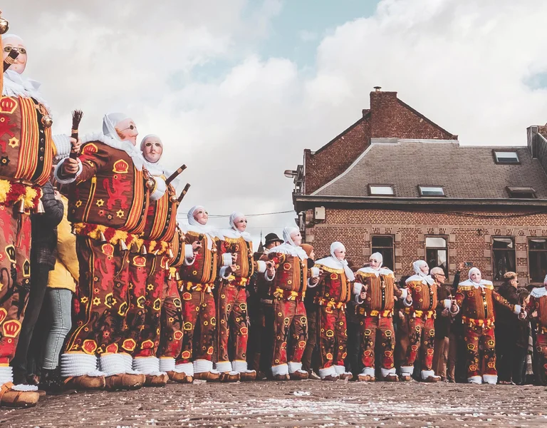 People in colorful costumes stand in a line at a town square with historic buildings.