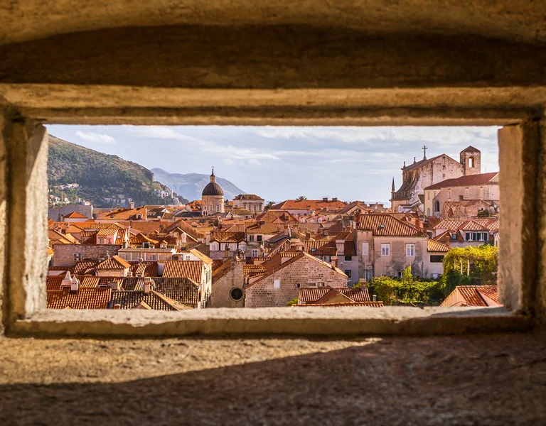Historic city with red rooftops viewed through a stone window, mountains in the background.
