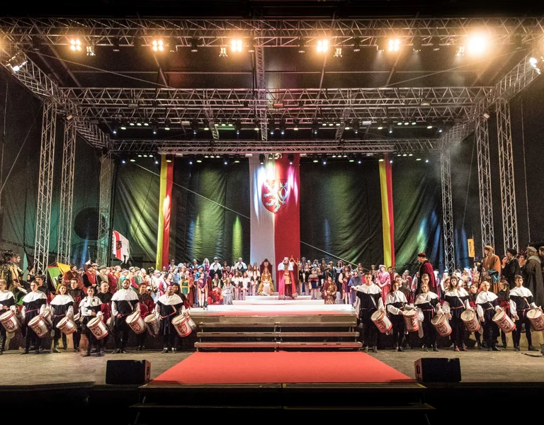 Large group in traditional costumes on a brightly lit stage with red carpet steps.
