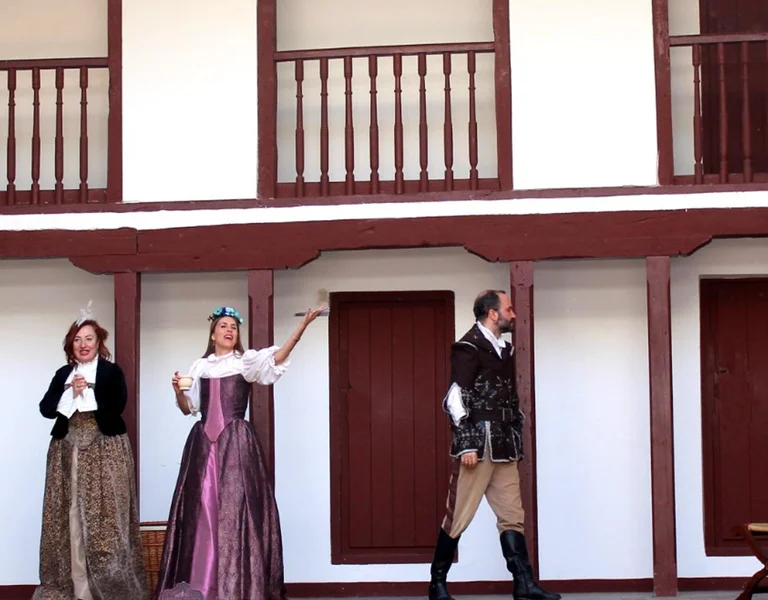 Three actors in period costumes perform on a wooden stage with balconies.