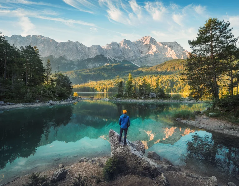 Person stands on rock by clear lake, surrounded by trees and distant mountains under blue sky.