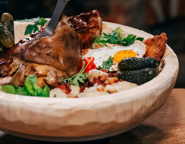 Bowl of roasted meat, fried egg, pickles, and greens on a rustic table.